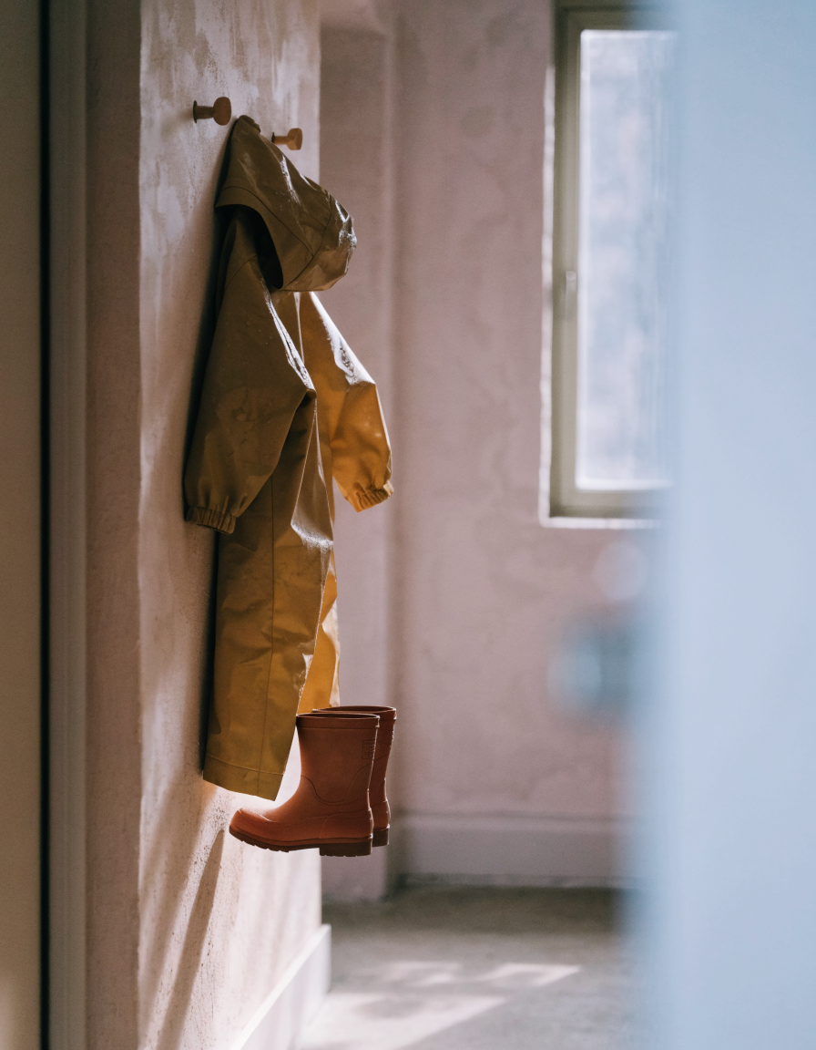 Child's mustard rain suit and terracotta rubber boots hanging on wooden pegs in a hallway