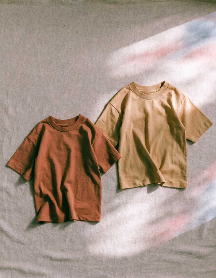 Two identical children's organic cotton t-shirts laid flat side by side on beige linen, one visibly smaller after washing