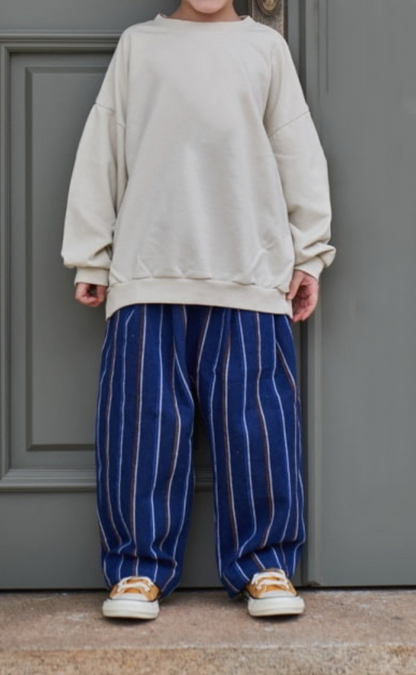 Child wearing oversized baggy striped denim pants with a beige sweater and sneakers, standing against a gray door background.